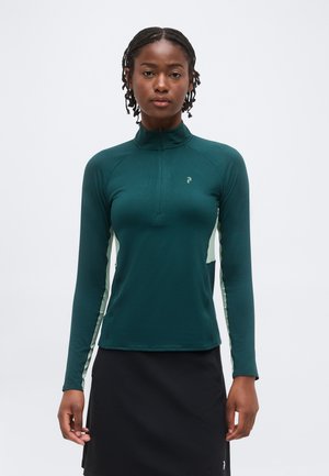 Femme aux cheveux tressés portant un haut bleu sarcelle foncé à manches longues avec une fermeture éclair et des panneaux latéraux clairs, associée à une jupe noire, debout devant un fond uni.