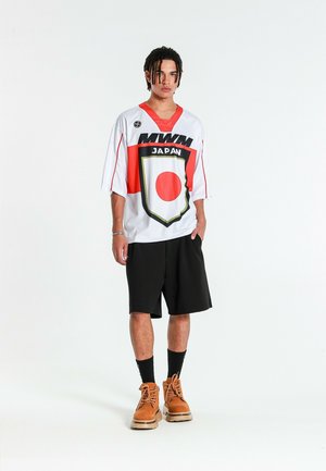 Joven con rastas lleva puesta una camiseta blanca y roja de gran tamaño con motivos de Japón, pantalones cortos negros, calcetines negros y botas de trabajo marrones sobre fondo blanco.