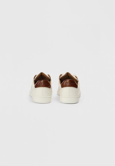 Baltas sporta kurpes ar gludu ādas augšdaļu, brūnas ādas papēža akcentiem un fakturētu gumijas zolēm. Papēdī ir iespiests logo.