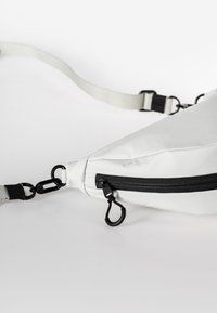 Weiße, dreieckige Tasche aus glattem Material, mit schwarzem Reißverschluss und Beschlägen, verstellbarem Trageriemen und minimalistischem Design.