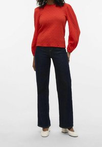 Rød tekstureret langærmet top med puffede ærmer, parret med mørkeblå brede bukser og hvide blokhælede sko.