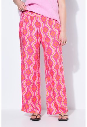 Osoba nosząca luźne różowe spodnie z falistym wzorem kwiatowym w kolorze pomarańczowym i żółtym, zestawiona z czarnymi klapkami i fakturowanym różowym topem.