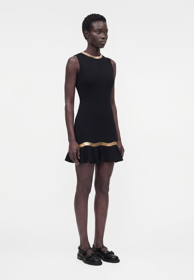 Γυναίκα που φοράει μαύρο αμάνικο φόρεμα με χρυσό περίγραμμα στον γιακά και το τελείωμα, σε συνδυασμό με μαύρα παπούτσια με αγκράφα, στέκεται σε λευκό φόντο.