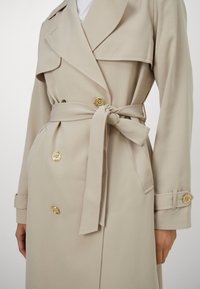 Trench-coat beige à double boutonnage avec boutons dorés, taille cintrée nouée en un nœud, et brides aux manches longues.