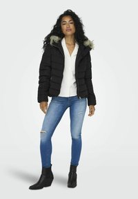 Svart puffjakke med fuskepelshette, quiltet design, glidelås, båret over en hvit skjorte, kombinert med slitte blå jeans og svarte ankelstøvler.