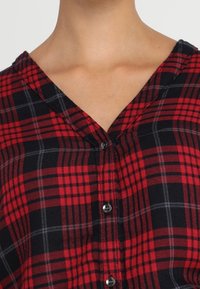 Chemise à carreaux rouge et noir à boutons, avec un col en V et des boutons noirs, en tissu doux et texturé avec un motif à carreaux.