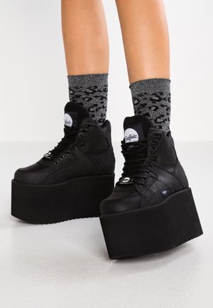 Nogi w czarnych sneakersach na platformie z grubymi podeszwami i czarno-szarych skarpetkach w panterkę, stojące na jasnej podłodze na białym tle.