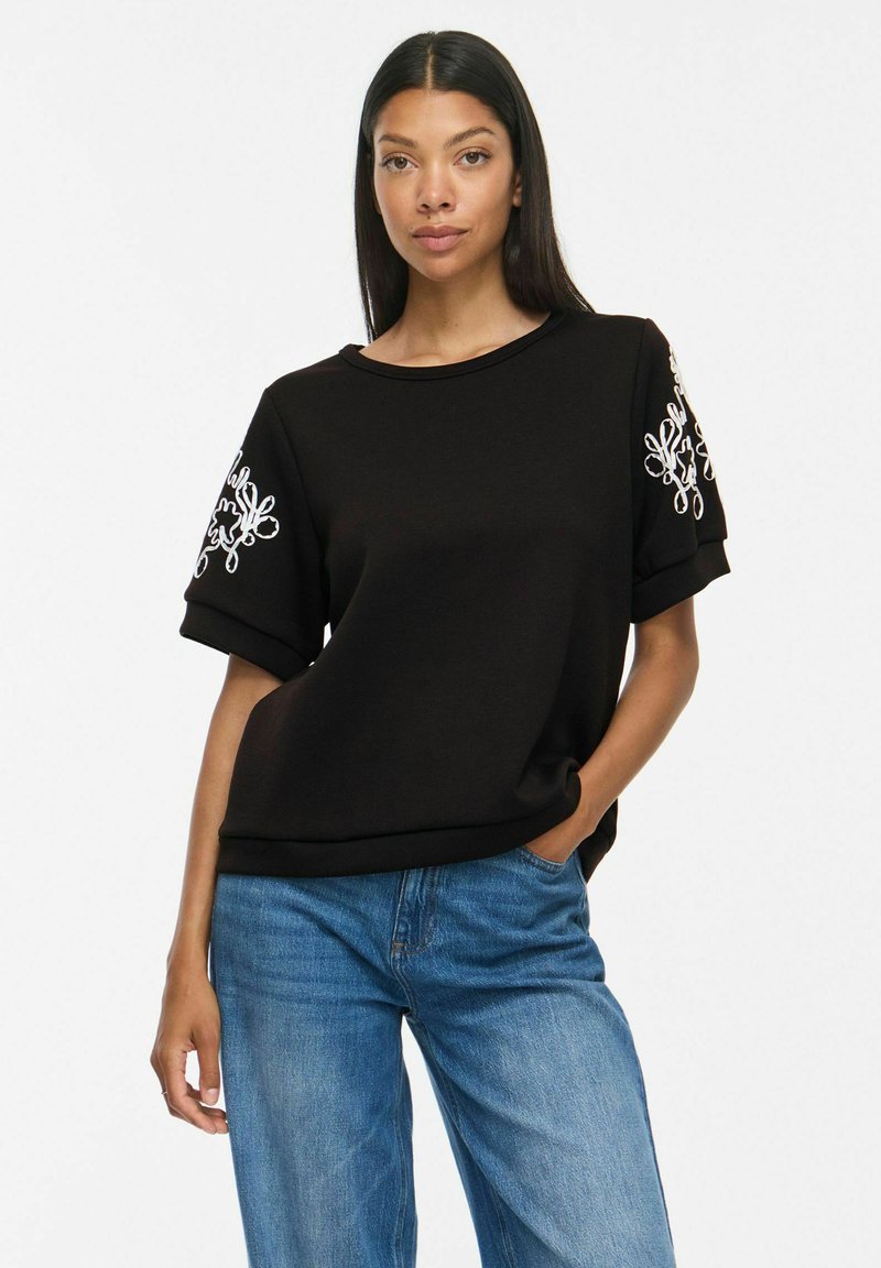 Femme aux longs cheveux foncés portant un haut noir à manches courtes avec une broderie blanche sur les manches et un jean bleu, debout avec une main dans la poche.