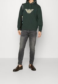 Mörkgrön huvtröja med en stor logotyp, i kombination med grå slim-fit jeans och mörkbruna tofflor. Inkluderar en svart väska.