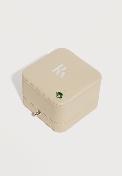 Bež nakitna škatla s srebrnim logotipom "RY" na vrhu, z zlatim uhanom z zelenim šestkotnim kamnom, ki počiva na pokrovčku.