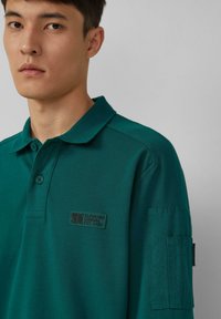 Türkises Polohemd mit Kragen, zwei Knöpfen und einem Aufnäher auf der Brust, der ein Logo zeigt. Weicher Stoff mit glatter Textur und taillierter Passform.