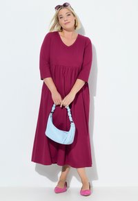 Švestkově zbarvené šaty s výstřihem do V a ¾ rukávy, s ozdobným pasem. Modelka drží světle modrou texturovanou kabelku a nosí růžové baleríny.