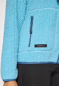 Lyseblå tekstureret fleecejakke med en lynlåslomme og sort label, båret over sorte bukser.