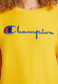 Felpa gialla caratterizzata da ricamo con il testo "Champion" in blu, con un accento logo rosso e blu sul lato sinistro del petto.
