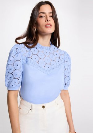 Frau trägt eine hellblaue Bluse mit Puffärmeln aus Lochspitze und cremefarbene High-Waist-Hose, posiert vor einem schlichten Hintergrund.