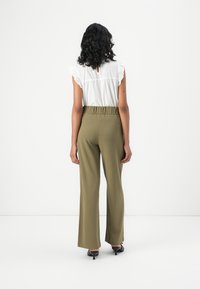 Blusa bianca con volant e dettaglio a chiave sul retro, abbinata a pantaloni ampi a vita alta di colore verde oliva e décolleté neri con punta affilata.