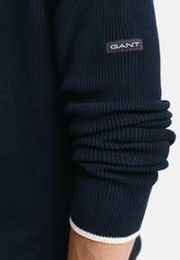 Granatowy sweter w prążki o grubej strukturze, z białym mankietem i małą naszywką logo GANT na górnej części rękawa.