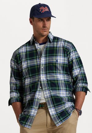 Geknoopte plaid shirt in groen, marineblauw en wit met opgerolde mouwen; gecombineerd met een marineblauwe pet met "Polo" in rode borduur. 