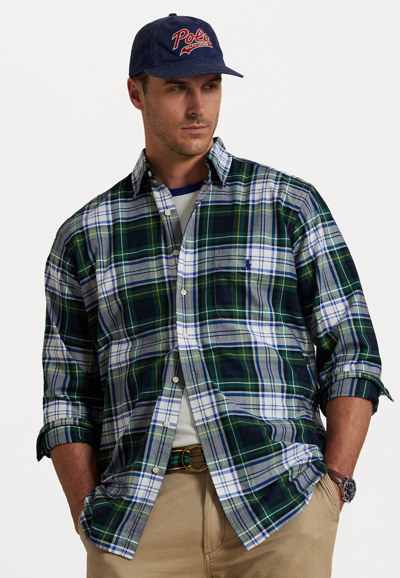 Geknoopte plaid shirt in groen, marineblauw en wit met opgerolde mouwen; gecombineerd met een marineblauwe pet met "Polo" in rode borduur. 