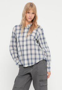 Camisa de botão em xadrez azul e branco, feita de tecido macio. Apresenta um corte descontraído com mangas longas e uma bainha arredondada. Adequada para uso casual.
