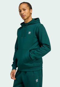 Zelená bavlnená mikina s kapucňou má predné vrecko, nastaviteľnú šnúrku a biele logo adidas na hrudi. Mäkká textúra s voľným strihom.