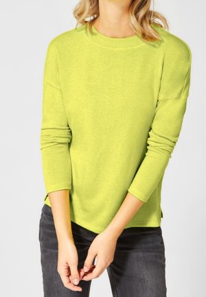 Pull over long en vert vif avec col rond côtelé, coupe décontractée et fentes latérales, associé à un jean gris foncé. Texture lisse.