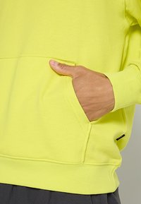 Hoodie vert citron en matériau doux et texturé, avec une poche avant et des poignets côtelés, arborant une petite étiquette de marque sur le côté.