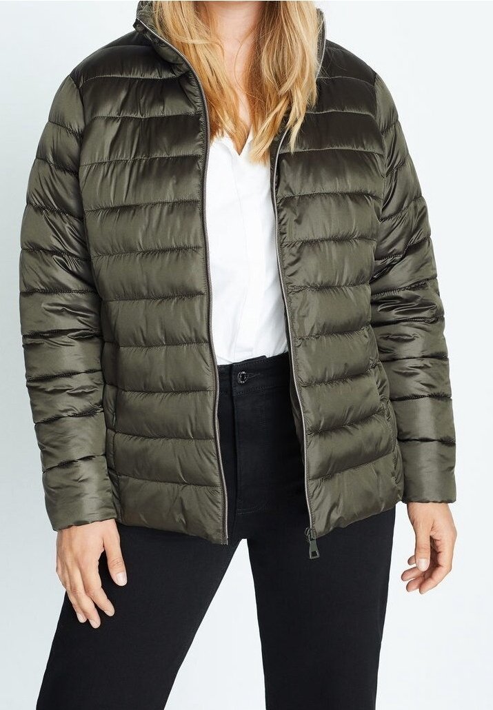 Chaqueta acolchada verde oliva con textura acolchada, cierre de cremallera, cuello alto y bolsillos laterales, llevada sobre una camisa blanca y pantalones negros.