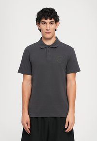 Mørkegrå polo-skjorte laget av bomullsblanding, med ribbestrikket krage, to knapper og en tonet logo-patch på venstre bryst.