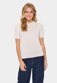 Svetle béžový krátký rukáv top s kulatým výstřihem a jemnými nabíranými rukávy, vyrobený z měkké, texturované látky. V kombinaci s tmavými džíny.