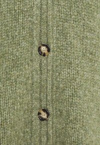 Green knitted cardigan featuring a textured pattern, two brown buttons, and visible stitches. Close-up view highlights material details.