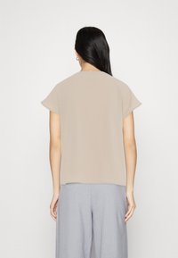 Donna con lunghi capelli scuri, che indossa una maglietta beige a maniche corte e pantaloni grigi larghi, in piedi fronte a uno sfondo chiaro e uniforme.