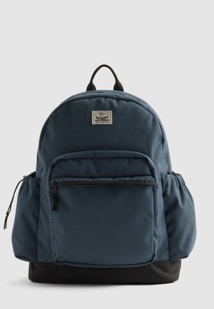 Mochila de tela azul marino con forma redondeada, que cuenta con un bolsillo frontal con cremallera, bolsillos de malla en los laterales y un fondo negro. Asas superiores resistentes.