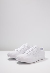 Baskets blanches avec une semelle en caoutchouc texturée, une tige synthétique lisse, et un logo étoile sur le côté, dotées d'un laçage frontal et d'un bout arrondi.