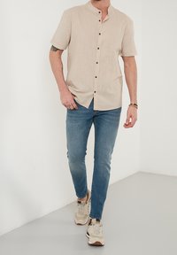 Beige linnen overhemd met korte mouwen en een mandarin kraag, gecombineerd met lichtblauwe slim-fit jeans en beige sneakers.