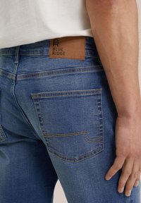 Jeans iz jeansa v srednji modri barvi s teksturiranim videzom, z rjavim usnjenim označevalcem "BLUE RIDGE" in rumenim šivanjem. Dva zadnja žepa sta vidna.