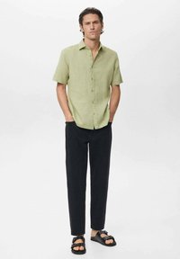 Camisa de botones de manga corta verde claro, pantalones negros ajustados y sandalias negras con hebilla. Textura suave, diseño informal.