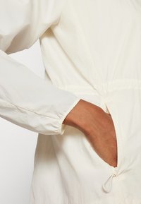Leichte, cremefarbene Jacke mit elastischen Bündchen und tailliertem Bund; verfügt über eine seitliche Tasche mit einer Kordelzug-Verschlussleiste.