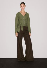 Grüne Leinen-Wickeloberteile kombiniert mit braunen weiten Hosen. Die Bluse hat lange Ärmel und ein gebundenes Taillendetail. Eine schlichte silberne Anhänger-Halskette ist inbegriffen.