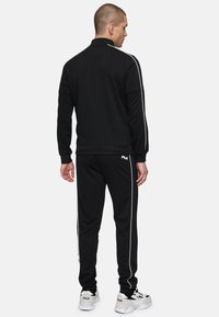 Chándal atlético negro con rayas laterales blancas, con un collar con cremallera y puños acanalados, usado con zapatillas blancas y negras.