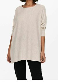 Femme portant un pull ample à manches longues de couleur beige clair avec un col rond et un pantalon noir, debout devant un fond blanc uni.
