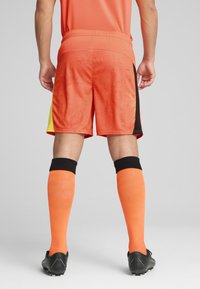 Pantalones cortos de fútbol naranjas con textura estampada, acentos laterales amarillos y ribetes negros; combinados con calcetas naranjas hasta la rodilla y botas negras.
