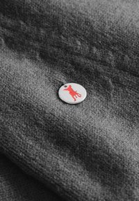 Szara tkanina o miękkiej teksturze, z okrągłym metalowym znaczkiem w kolorze srebrnym, na którym znajduje się sylwetka psa w kolorze czerwonym.