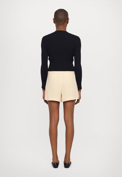 Crna rebrasta pulover, uskog kroja, s dugim rukavima. Bež kratke hlače viskog struka, krojene s stražnjim džepovima. Crne balerinke.