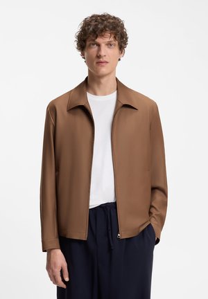 Giovane uomo con capelli ricci che indossa una giacca marrone con zip sopra una camicia bianca e pantaloni blu scuro, in piedi su uno sfondo bianco.