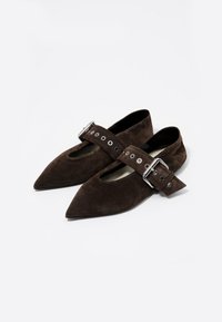 Zapatos planos de ante marrón con punta afilada, que presentan una amplia correa de tobillo con hebilla metálica y recortes circulares a lo largo de la correa.