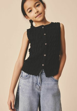 Młoda dziewczyna z warkoczami, ubrana w bezrękawnik na guziki w kolorze czarnym oraz luźne, jasnoniebieskie dżinsy, stojąca na beżowym tle.