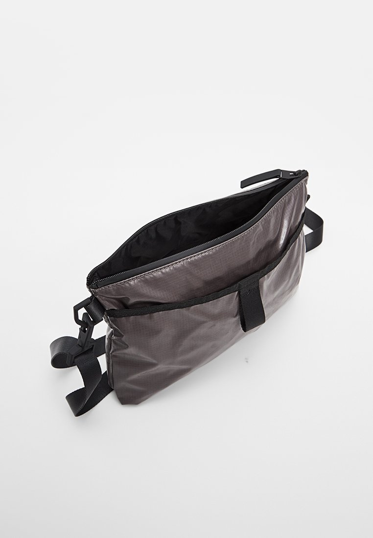 Kleine graue Umhängetasche mit schwarzem verstellbarem Trageriemen, teilweise geöffneter Reißverschluss und Vordertasche mit Klettverschluss, vor weißem Hintergrund gezeigt.