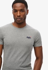 Grå T-shirt med korte ærmer lavet af tekstureret stof. Har et lille blåt logo på venstre brystområde. Enkel, tætsiddende design.