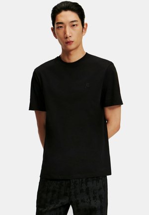 Ung mand med sort t-shirt med korte ærmer og mørke, mønstrede bukser står mod en ensartet lys baggrund.